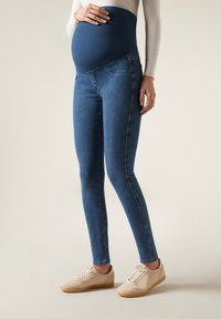 Jeans de maternidad con un panel superior azul elástico y una pierna de mezclilla ajustada. Presenta bolsillos delanteros y una textura suave, combinados con zapatillas de color claro.