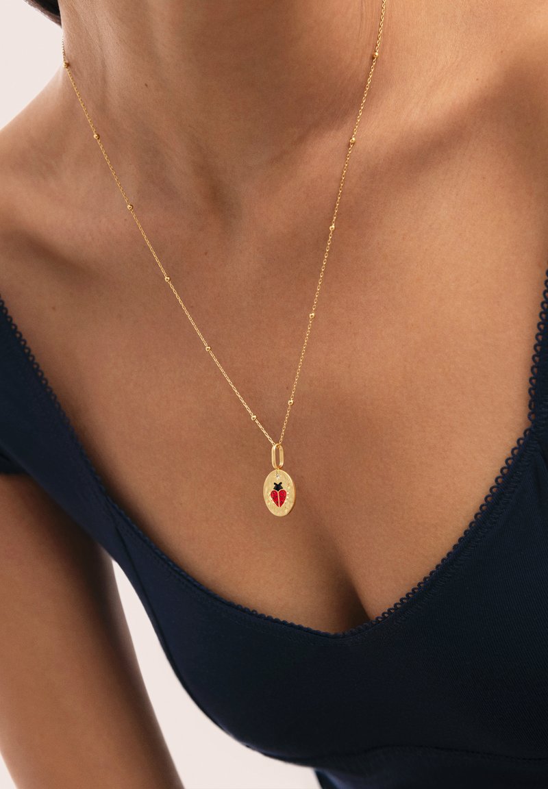 Collar de oro con colgante ovalado que presenta una mariquita roja y negra, llevado sobre una blusa azul oscuro con escote en V en el cuello y pecho de una mujer.