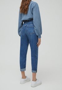 Jeans en denim bleu taille haute, légèrement fuselés avec des revers retroussés, dotés de poches arrière et d'une finition texturée. Portés avec des baskets blanches.