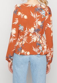 Blumenbluse mit rostfarbenem Hintergrund, langen Ärmeln, tailliertem Schnitt und blauen sowie cremefarbenen Blumenmustern. Kombiniert mit hellblauen Jeans.