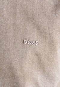 Una tela color arena con una textura suave presenta la palabra "BOSS" bordada en blanco, ligeramente desplazada hacia la derecha.