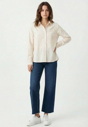Femme aux cheveux bruns ondulés portant une chemise beige à boutons avec un motif subtil, un jean bleu à jambes larges et des baskets blanches, debout avec une main sur la hanche.