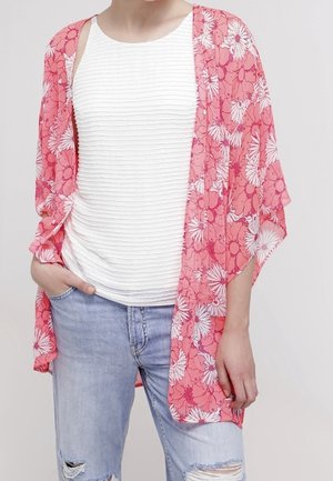 Person trägt ein weißes strukturiertes Tanktop, hellblaue zerrissene Jeans und einen rosa floralen offenen Cardigan.