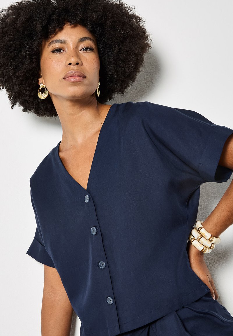 Femme avec une coiffure afro naturelle portant des boucles d'oreilles créoles en or, un haut bleu marine à manches courtes boutonné et des bracelets blancs épais au poignet.