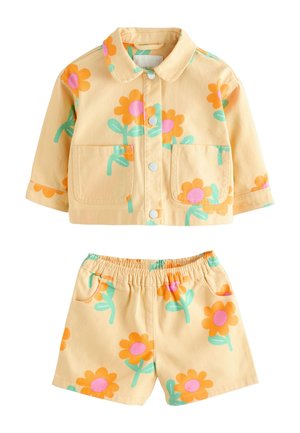 Maluch z warkoczami nosi żółtą kurtkę w kwiaty i pasujące krótkie spodenki, stoi w różowych sandałach na beżowym tle.