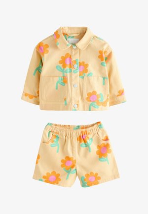 Småbarn med flettet hår iført gul blomsterjakke og matchende shorts, stående med lyserøde sandaler mod beige baggrund.
