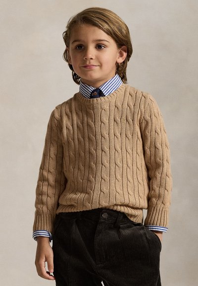 Bambino con capelli castano chiaro indossa un maglione a trecce color sabbia sopra una camicia a righe blu e pantaloni di velluto a coste neri, in piedi con una mano in tasca.