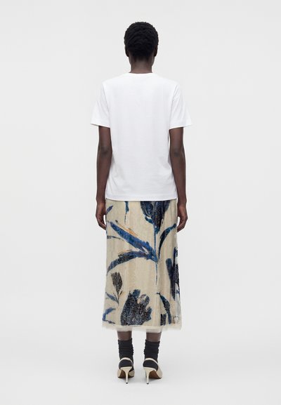 Personne debout, dos à la caméra, portant une chemise blanche à manches courtes, une jupe midi fleurie, des chaussettes noires et des talons hauts beiges sur un fond uni.