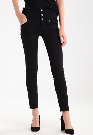 Kvinde iført sorte højtaljede skinny jeans med tre knapper foran, sorte hæle, stående med den ene hånd i lommen.
