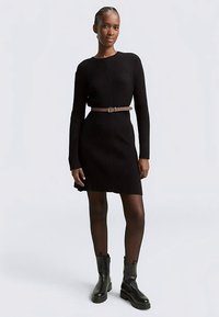 Czarna prążkowana sukienka z dzianiny z długimi rękawami, okrągłym dekoltem i dopasowaną talią, zestawiona z jasnobrązowym paskiem i czarnymi botkami.