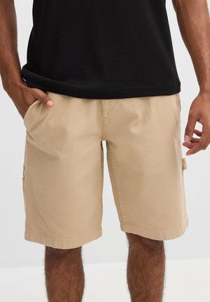 Hombre vistiendo pantalones cortos beige hasta la rodilla y una camiseta negra, con una mano en el bolsillo y la otra relajada a su lado.