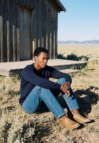 Homme en pull bleu, jean et bottes marron assis par terre avec les genoux relevés, près d'une cabane en bois dans un paysage sec et ouvert sous un ciel dégagé.