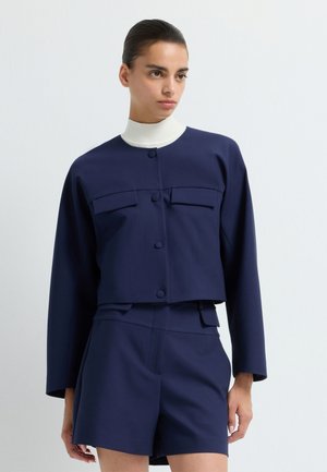 Marineblå cropped jakke med to brystlommer og knaplukning, kombineret med matchende shorts, med en hvid ribbet krave.