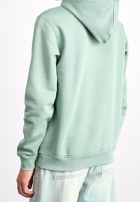 Mintgrön hoodie med mjuk textur, kengurufickor och justerbar dragsko på huvan. Sammanställd med ljus tvättade denimbyxor.