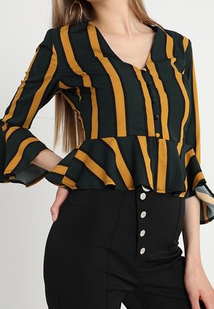Kobieta ubrana w czarno-musztardowy prążkowany top peplum z rozkloszowanymi rękawami oraz czarne, wysoki stan spodnie z guzikami z przodu.