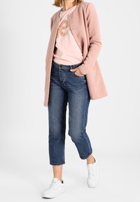 Ljusrosa texturerad cardigan över en rosa t-shirt med en logotyp, i kombination med mörkblå korta jeans och vita sneakers.