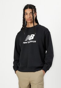 Črna aktivna majica s krožnim izrezom, na kateri je vidna bela "New Balance" oznaka čez prsni del. Dolgi rokavi, teksturirana tkanina.