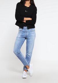 Cardigan noir texturé avec encolure ronde et boutons, associé à un jean en denim bleu clair coupe droite avec ourlet effiloché, complété par des baskets blanches.