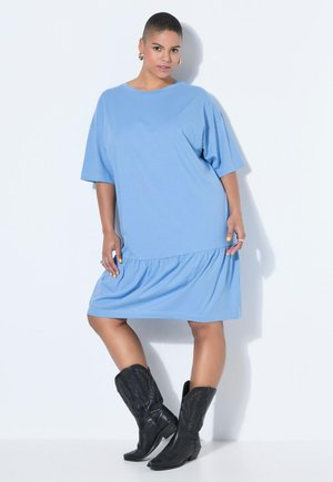 Mujer con el cabello corto que lleva un vestido suelto azul claro hasta la rodilla y botas de vaquero negras, de pie contra un fondo blanco.