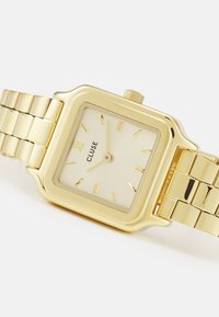Montre-bracelet rectangulaire en or avec un bracelet en or texturé, dotée d'un cadran crème avec des index horaires dorés et d'une petite couronne en haut.