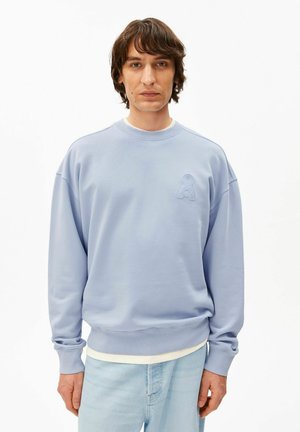 Homme portant un sweat-shirt bleu clair avec un petit logo texturé sur la poitrine, associé à un jean bleu clair, debout devant un fond blanc.