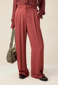 Vrouw in rustkleurige wijde broek en bordeauxrode blouse, met een tas van luipaardprint, en zwarte schoenen op een neutrale achtergrond.