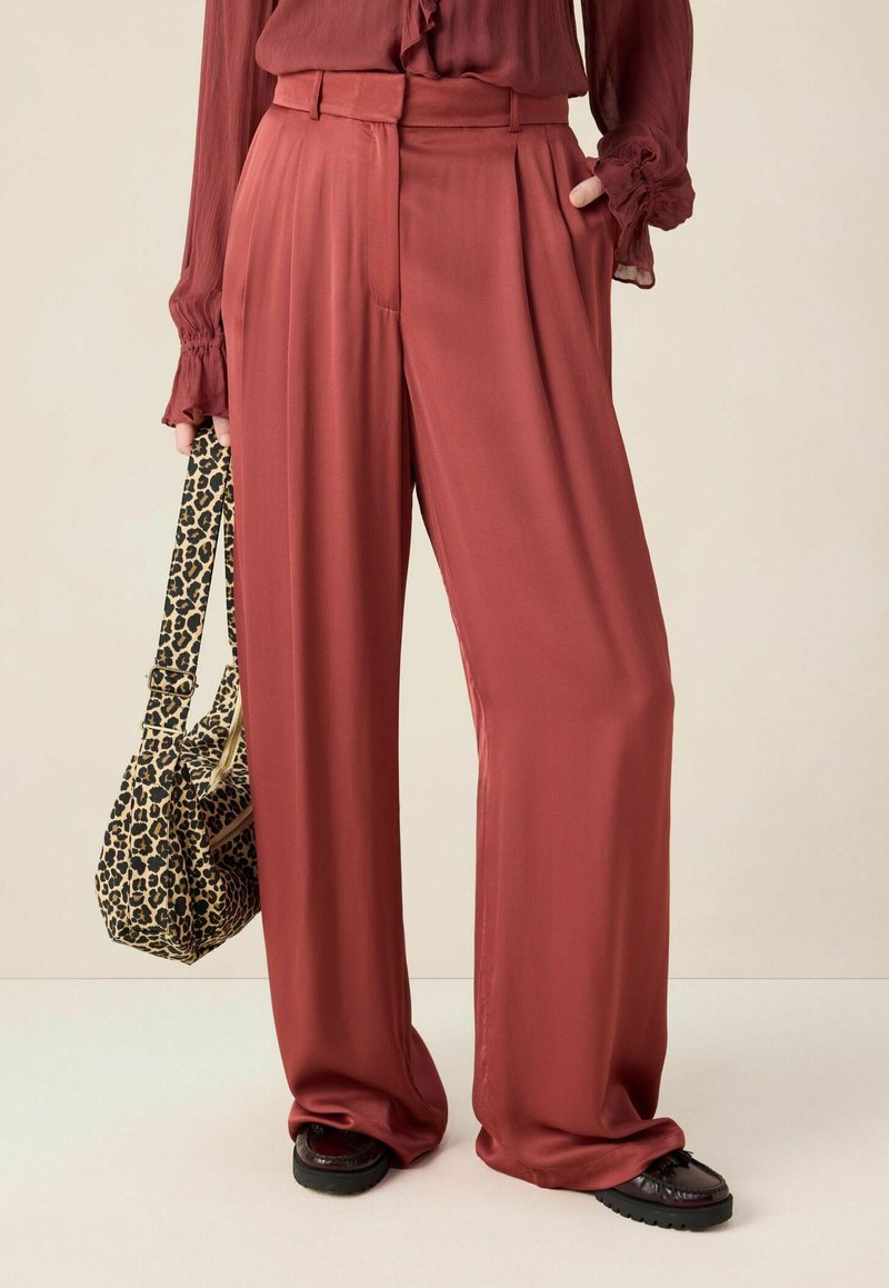 Vrouw in rustkleurige wijde broek en bordeauxrode blouse, met een tas van luipaardprint, en zwarte schoenen op een neutrale achtergrond.