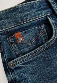 Nahaufnahme einer blauen Denim-Jeans-Tasche mit oranger Naht, einem kleinen braunen Lederetikett und Metallnieten.