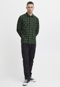 Cămașă flanelă verde cu model tartan, cu o rețea albă, cu închidere cu nasturi pe față, două buzunare la piept; asociată cu pantaloni închiși la culoare și adidași negri.