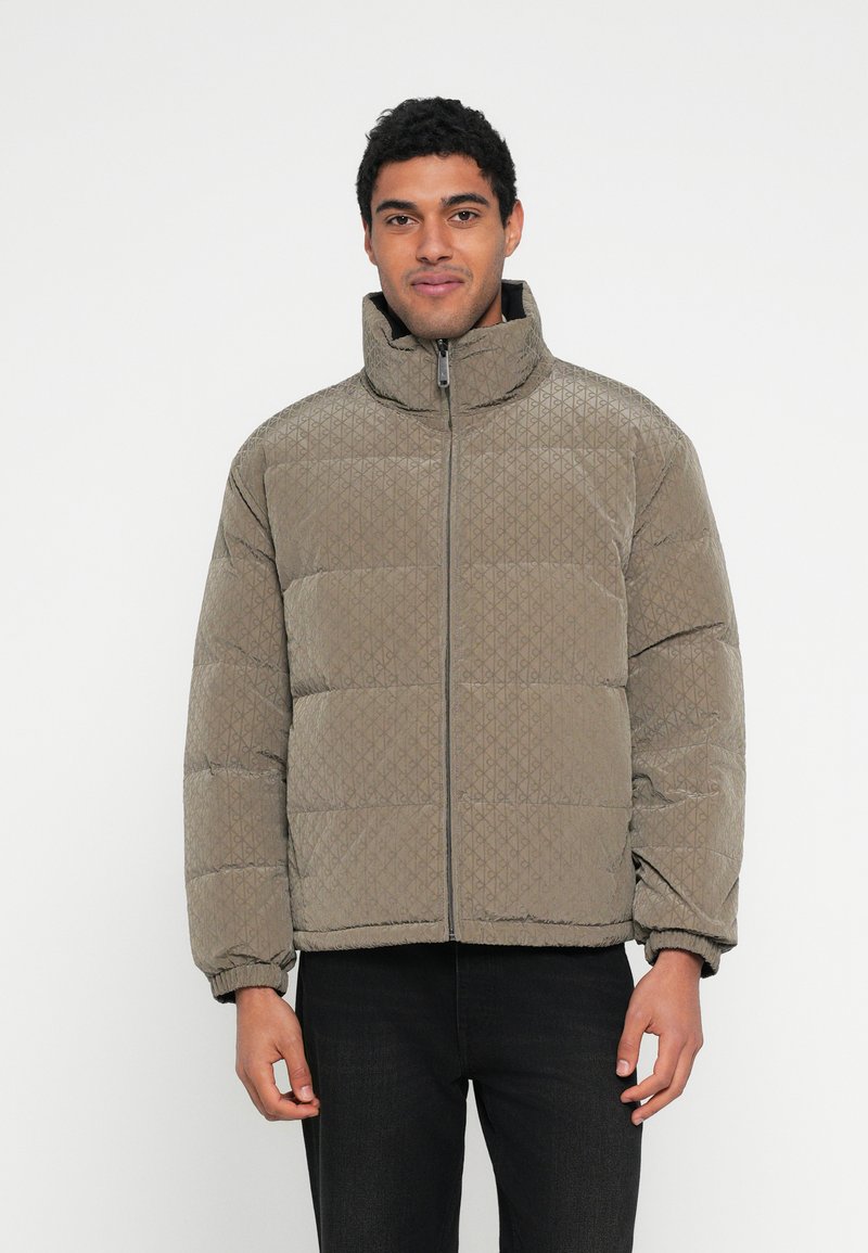 Geacă puf maro cu guler înalt, model matlasat, fermoar frontal și manșete elastice, purtată peste pantaloni negri.