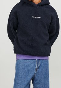 Granatowa bluza z polaru o fakturze przypominającej owczą wełnę, z białym logo "Original Studio" z przodu oraz luźnym krojem.