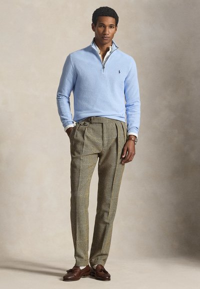 Mann trägt hellblauen Viertel-Zip-Pullover, weißes Hemd mit Kragen, gemusterte hellbraune Hose und braune Tasselloafers, steht mit Händen in den Taschen.
