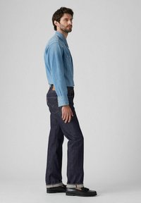 Camisa de mezclilla azul claro con cierres de botones, combinada con jeans azul oscuro con puños enrollados y un parche de cuero marrón. Mocasines negros complementan el look.