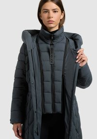 Napihnjena siva bunda z visokim ovratnikom in sprednjim zadrgo. Ima vodoravne šive in snemljivo notranjo plast. Gladek teksturiran, lahka zasnova.