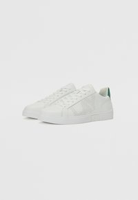 Weiße Ledersneaker mit durchbrochenem Design, flacher weißer Gummisohle, grüner Akzent an der Ferse und strukturiertem Branding an der Seite.
