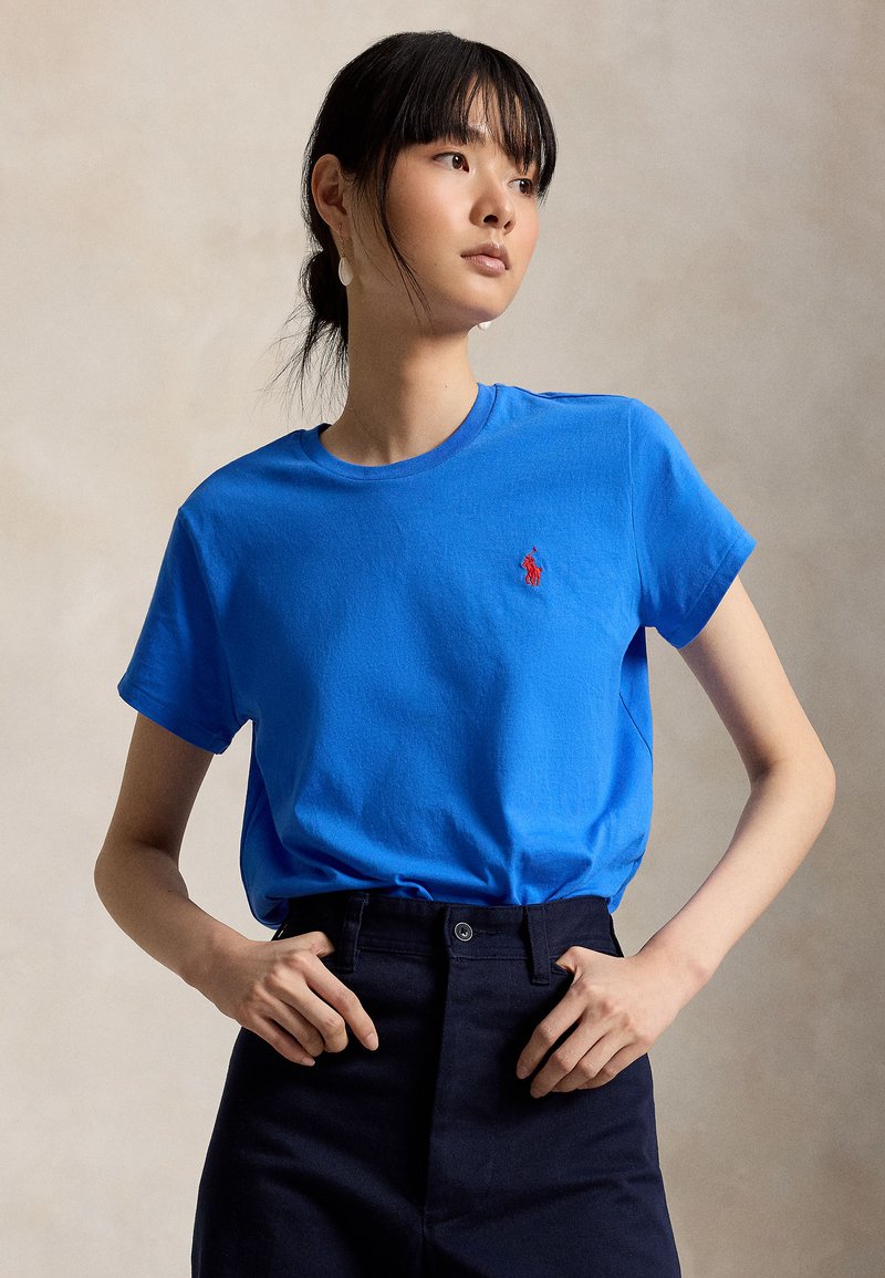 Frau mit dunklen Haaren in einem blauen T-Shirt mit rotem Logo, trägt schwarze Hose, steht mit den Händen in der Taille vor neutralem Hintergrund.