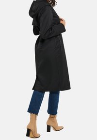 Frau in einem schwarzen, kapuzenbewehrten Trenchcoat, mit verkürzten blauen Jeans und beigen Wildleder-Stiefeletten mit Absatz, die im Profil steht.