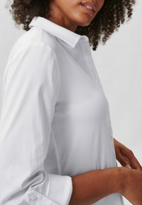 Witte langemouwen shirt met een klassieke kraag, met een gladde textuur en knoopsluiting bij de manchetten. Weergegeven onder een lichte hoek.