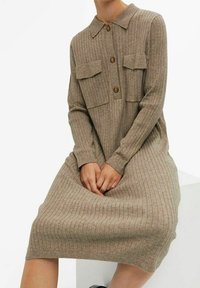 Robe en maille côtelée beige mi-longue avec manches longues, deux poches à rabat sur la poitrine, un col et trois boutons sur le devant.