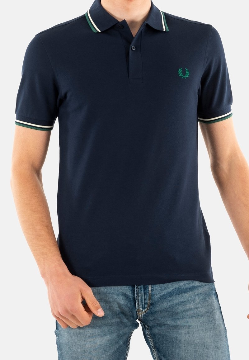 Homme portant un polo bleu marine avec un col et des manches rayés verts et blancs, associé à un jean bleu clair.