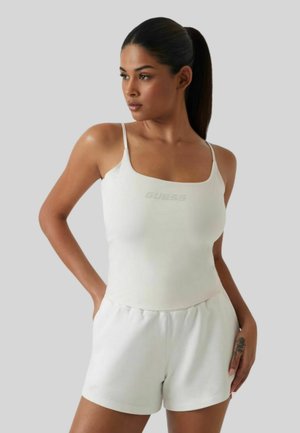 Donna con coda di cavallo che indossa un top bianco con spalline sottili e pantaloncini bianchi, posando con le mani nelle tasche su uno sfondo semplice.