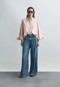 Ροζ ανοιχτόχρωμο πουκάμισο με κουμπιά και μανίκια γυρισμένα, συνδυασμένο με φαρδιά ξεβαμμένα μπλε τζιν. Το μοντέλο φορά καφέ γυαλιά ηλίου. Απλός, σύγχρονος στυλ.