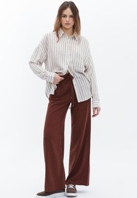 Camicia bianca oversize a righe; pantaloni larghi color ruggine; modella che indossa sneakers, mani in tasca. Styling semplice e strutturato.