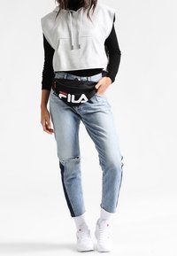 Sweat à capuche gris court porté sur un haut à manches longues noir, associé à un jean bleu clair délavé avec des bandes latérales bleu marine et des baskets blanches.