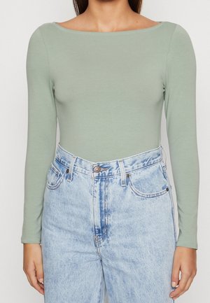 Vrouw draagt een lichtgroen nauwsluitend shirt met lange mouwen, ingestopt in een hooggewaisterde lichtblauwe gewassen spijkerbroek.