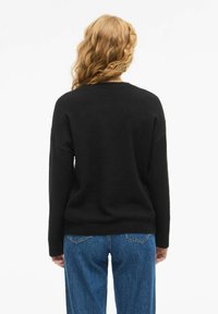 Person med långt lockigt hår som bär en svart tröja och blå jeans, vänd bortåt mot en helt vit bakgrund.