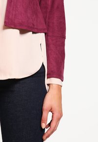 Burgundy faux suède-jacka med asymmetrisk nederkant, lager på en ljusrosa blus. Mörka jeans delvis synliga. Hand på jackan.