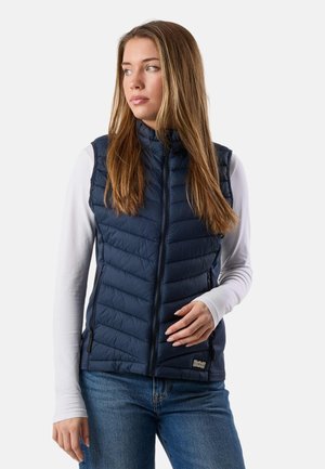 Kvinde med langt hår iført en marineblå quiltet ærmeløs vest over en hvid langærmet skjorte og blå jeans, kiggende mod venstre.