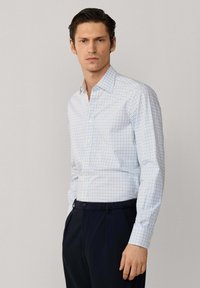 Homme portant une chemise blanche cintrée à carreaux bleus et verts, associée à un pantalon bleu marine foncé, debout devant un fond uni.
