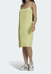 Kobieta w jasnozielonej, bezrękawnikowej sukience midi z cienkimi ramiączkami i białych trampkach z czarnymi paskami.